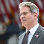 Scott Bessent in a suit with an American flag pin stands outdoors, with blurred U.S. flags and a presidential podium visible in the background.