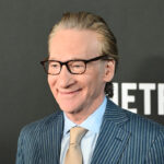 Bill Maher smiles in front of a black backing with white writing that reads "Netflix."