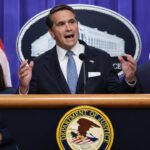 A man in a suit speaks at a podium with the Department of Justice seal, flanked by a woman and another man, both in business attire. American flags and an official emblem are visible in the background.