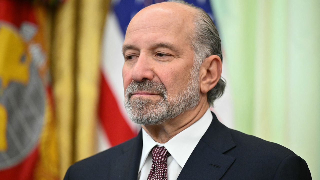 Home 1 A man with a beard and mustache, wearing a dark suit, white shirt, and patterned tie, stands in front of blurred flags.