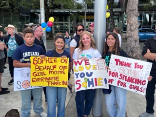 Why These Women Rallied for Sophie Cunningham in Seattle 1 Four people stand outdoors holding colorful signs that thank Sophie Cunningham for advocating for girls. There are more people and trees in the background, with balloons attached to a pole.