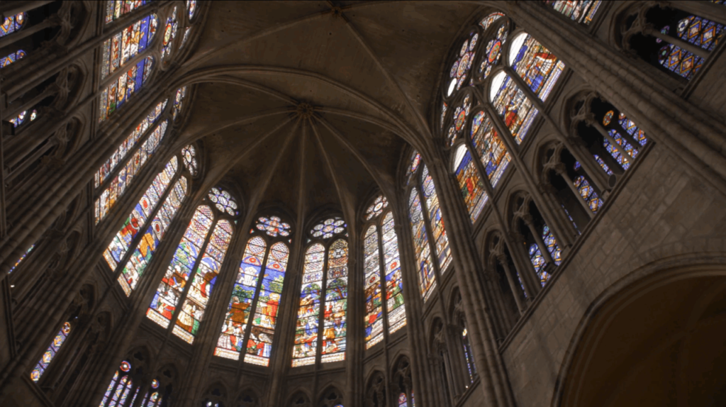 Bishop Barron’s First Theatrical Film Isn’t Nostalgia. It’s a Challenge. 2 Interior view of a Gothic cathedral with high vaulted ceilings and large, colorful stained glass windows depicting religious scenes. Light filters through the windows, illuminating the stone architecture.
