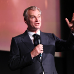 A man in a suit speaks into a microphone on stage, gesturing with one hand. Another man in a suit stands blurred in the background.