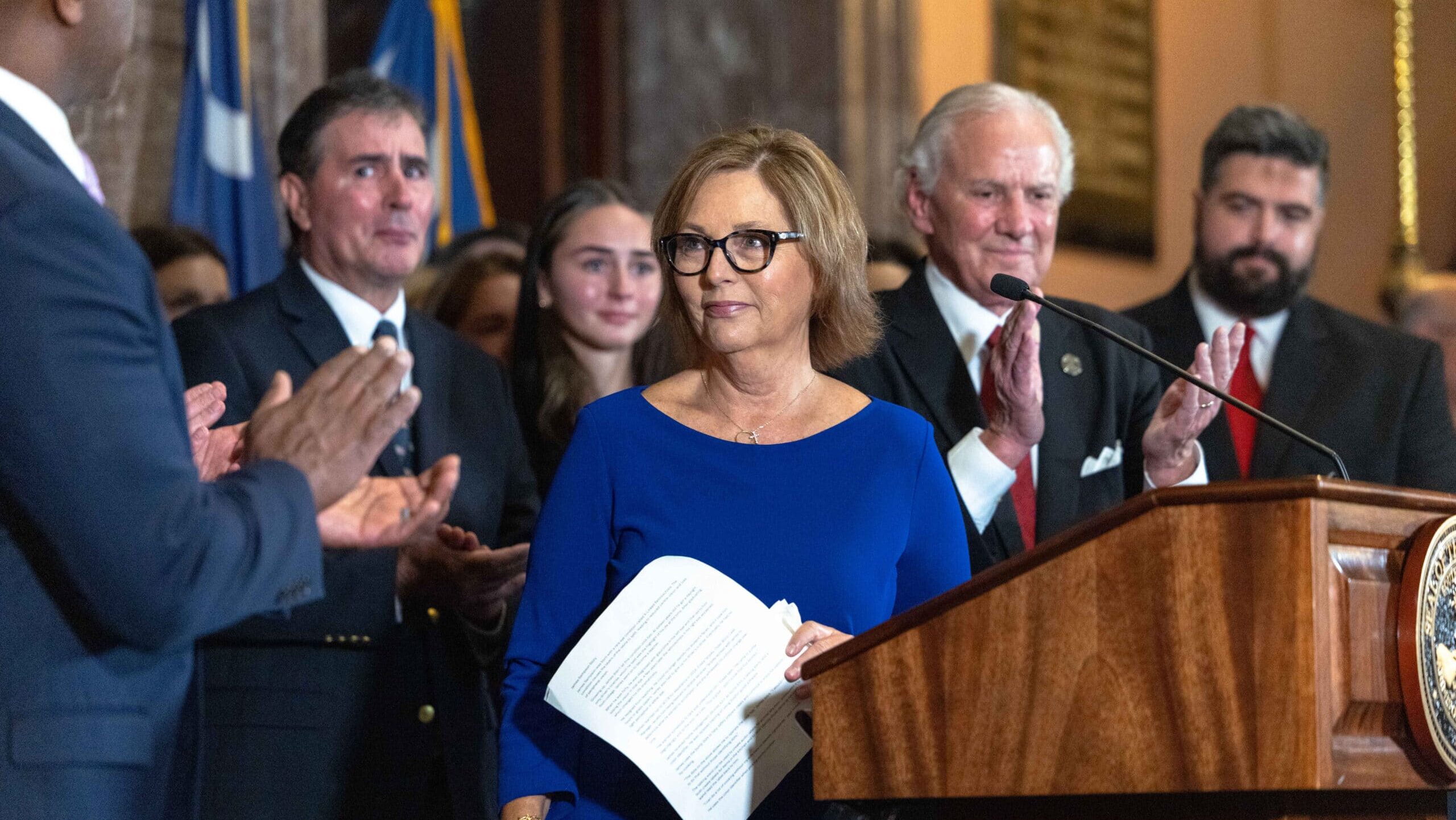 Darline Graham Nordone stands at a podium, surrounded by people applauding her in a formal indoor setting with flags in the background.