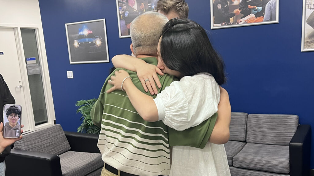 Americans Are Embracing Communism. Pastor Ezra Jin Felt Its Iron Fist. 1 Three people embrace in a group hug in a room with blue walls and framed photos. One person holds a phone displaying a photo of someone—perhaps Jin. There is a gray couch and a potted plant in the background.