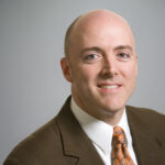 A man in a brown suit, white shirt, and orange patterned tie smiles at the camera against a plain light gray background.