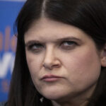 Haley Stevens, with a serious expression is shown in close-up against a blue background with partially visible white and orange fund text.