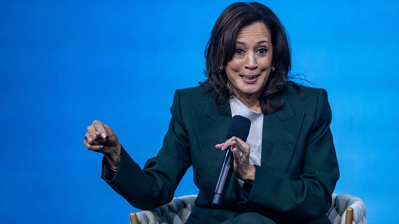 A woman in a dark green suit sits on a chair, holding a microphone and gesturing with her left hand, speaking in front of a blue background.