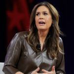 Gov. Sanders sits speaking on stage, wearing a brown leather top. Against a background featuring blurred red, white, and blue colors and shapes, she passionately discusses the importance of education.