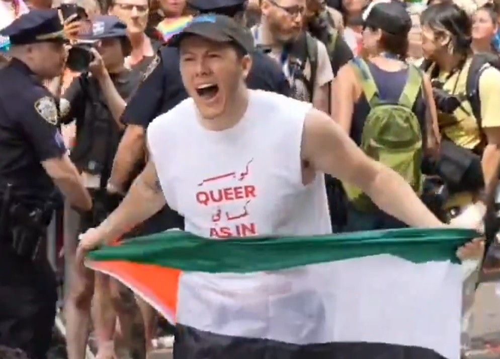Time to Wave ‘Queer’ Goodbye 1 A person in a crowd holds a Palestinian flag and shouts, wearing a shirt that reads Queer as in in English and Arabic. Police officers and other protesters are visible in the background.