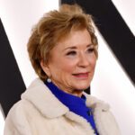 Linda McMahon with short light brown hair, wearing a blue top and a white fur coat, smiles in front of a white background with large black letters that spell out "Nation.