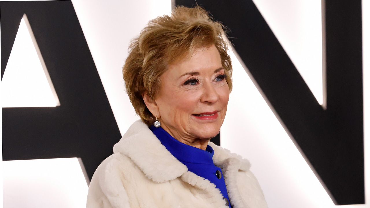 Linda McMahon with short light brown hair, wearing a blue top and a white fur coat, smiles in front of a white background with large black letters that spell out "Nation.