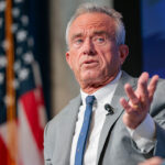 A man in a gray suit gestures while speaking onstage, with an American flag and a blue backdrop visible behind him.