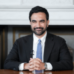 Zohran Mamdani sits at a table. He wears a small microphone on his lapel. The background features a fireplace with a white mantel, evoking the classic charm of Little Italy.