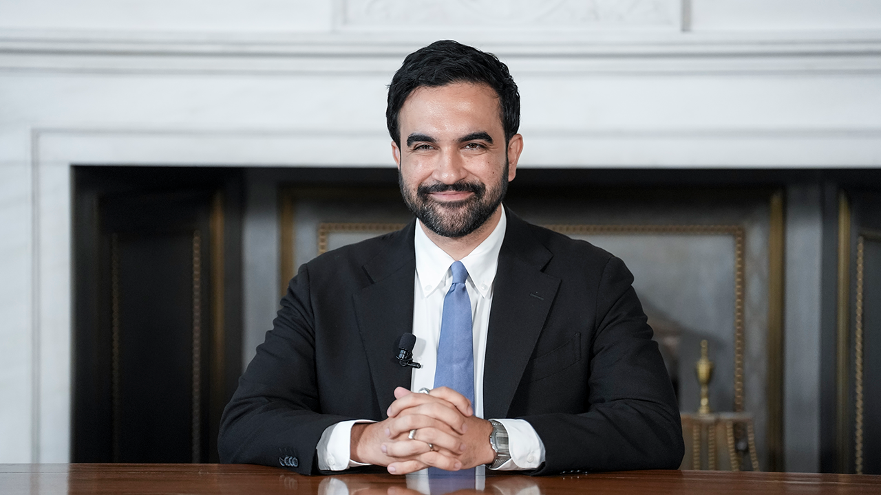 Home 1 Zohran Mamdani sits at a table. He wears a small microphone on his lapel. The background features a fireplace with a white mantel, evoking the classic charm of Little Italy.