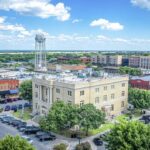 Aerial view of a small city square with a historic courthouse, surrounding trees, parked cars, brick buildings, and a water tower under a blue sky with scattered clouds.