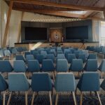 Rows of blue chairs face a stage with two large screens and a podium in a modern, well-lit auditorium with wooden beams and large windows.