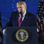 Trump stands at a podium with the presidential seal, speaking into a microphone about the dangers of communism. Multiple American flags are displayed behind him against a blue backdrop.