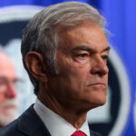 A serious-looking man in a suit and red tie stands in focus, with another man blurred in the background. A circular emblem and blue curtain are visible behind them.