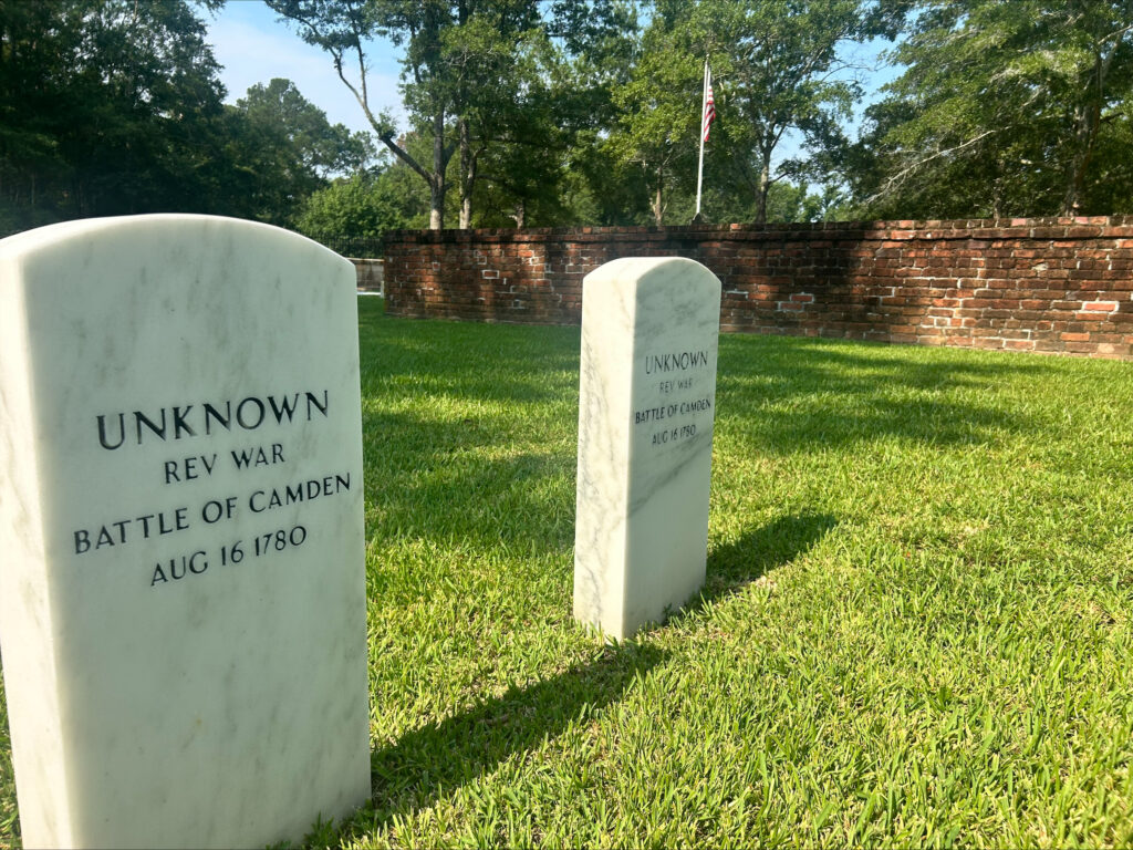 Rest Well, Young Patriot: Archeological Discoveries From the Battle of Camden 1 Two white gravestones read UNKNOWN REV WAR BATTLE OF CAMDEN AUG 16 1780, placed on green grass near a brick wall and trees, with an American flag in the background, marking the historic site of the Battle of Camden.