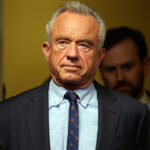A man in a suit and tie stands indoors with a serious expression, facing forward. There is another person blurred in the background and the setting appears to be an office or hallway.