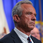 A man in a suit stands in front of blue and white flags, looking serious. The background is blurred, and he is facing left with a focused expression.