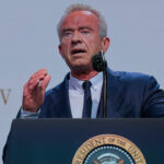 A man in a blue suit speaks at a podium with the U.S. presidential seal, gesturing with his right hand. The background shows partial text ending in Rx.gov..