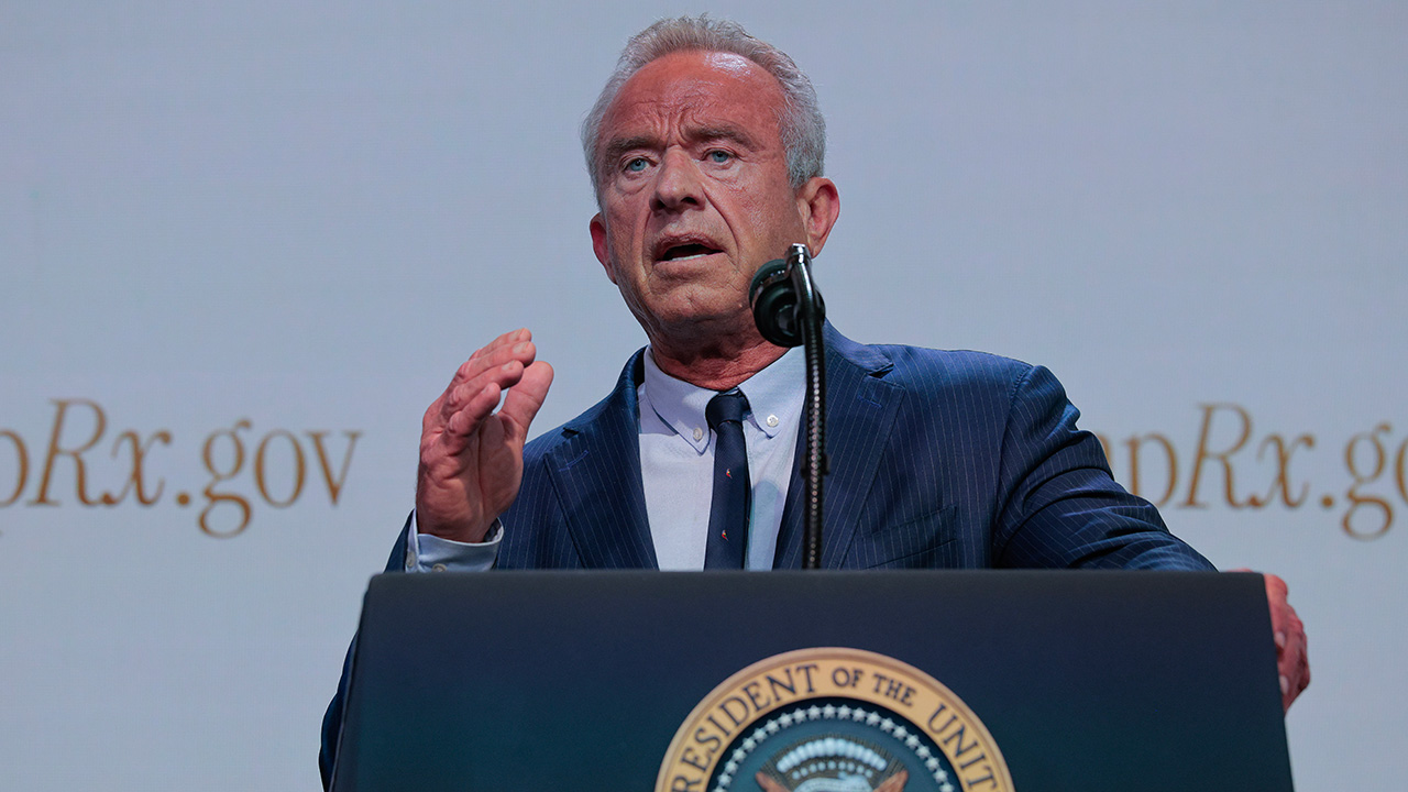 A man in a blue suit speaks at a podium with the U.S. presidential seal, gesturing with his right hand. The background shows partial text ending in Rx.gov..