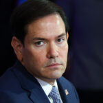 A man in a navy blue suit and striped tie sits in a chair, looking slightly to the side with a serious expression. An American flag pin is visible on his lapel. The background is blurred.