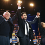 Three people on stage hold hands and raise them in celebration before a cheering crowd. Many in the audience hold blue and yellow signs that read Zohran. The atmosphere is energetic and festive.