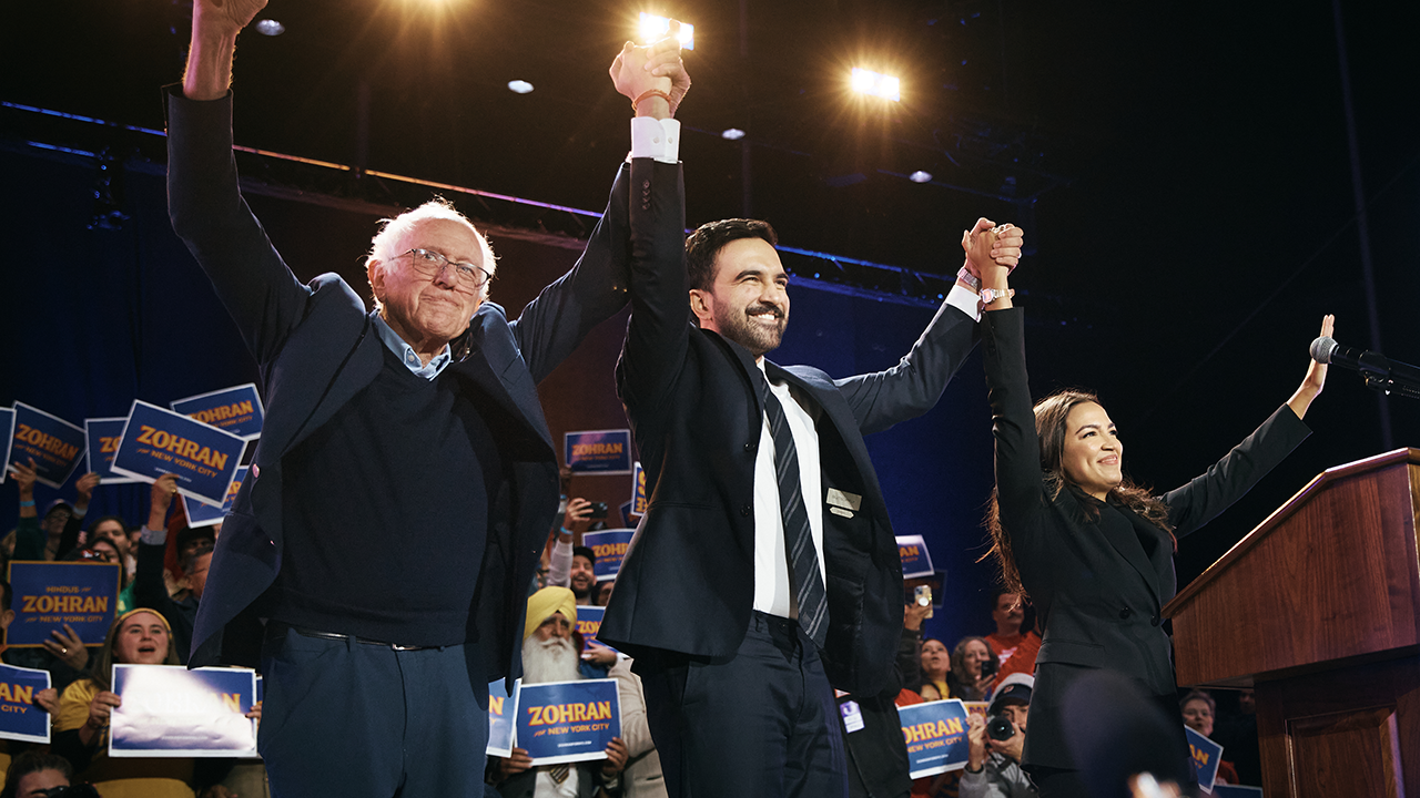 Home 2 Three people on stage hold hands and raise them in celebration before a cheering crowd. Many in the audience hold blue and yellow signs that read Zohran. The atmosphere is energetic and festive.