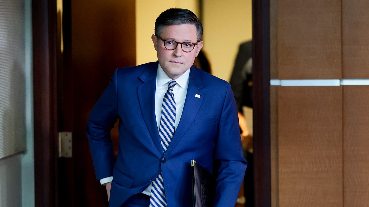 A man in a blue suit and striped tie walks through a doorway holding a folder, with a neutral expression, in a professional office setting.