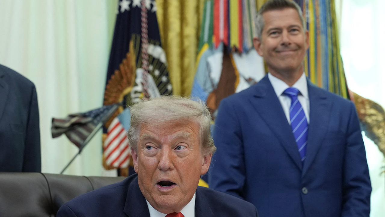 A man with blond hair sits at a desk, looking surprised, while another man in a blue suit stands behind him smiling. Flags and a gold curtain are visible in the background.