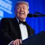 A man in a black tuxedo smiles while speaking at a podium with a microphone. A blue background with white text is visible behind him.