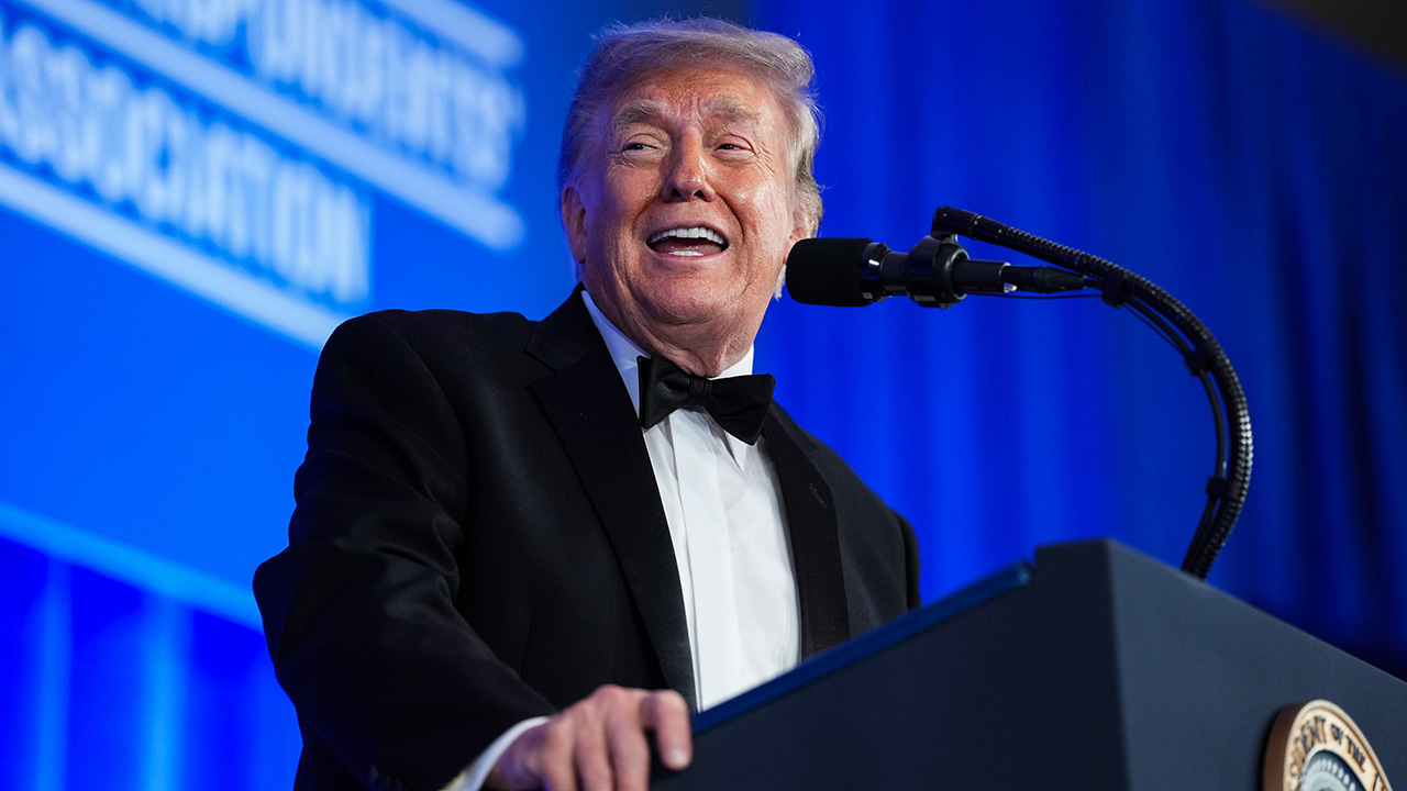 A man in a black tuxedo smiles while speaking at a podium with a microphone. A blue background with white text is visible behind him.