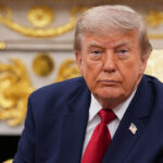 A man in a navy suit with a red tie and U.S. flag pin sits in an ornate room with gold decorations, looking forward with a serious expression.