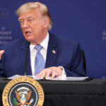 A man in a blue suit speaks at a table with a presidential seal, microphone, and water bottle, against a blue backdrop with Raterpayer Protection Pledge text.